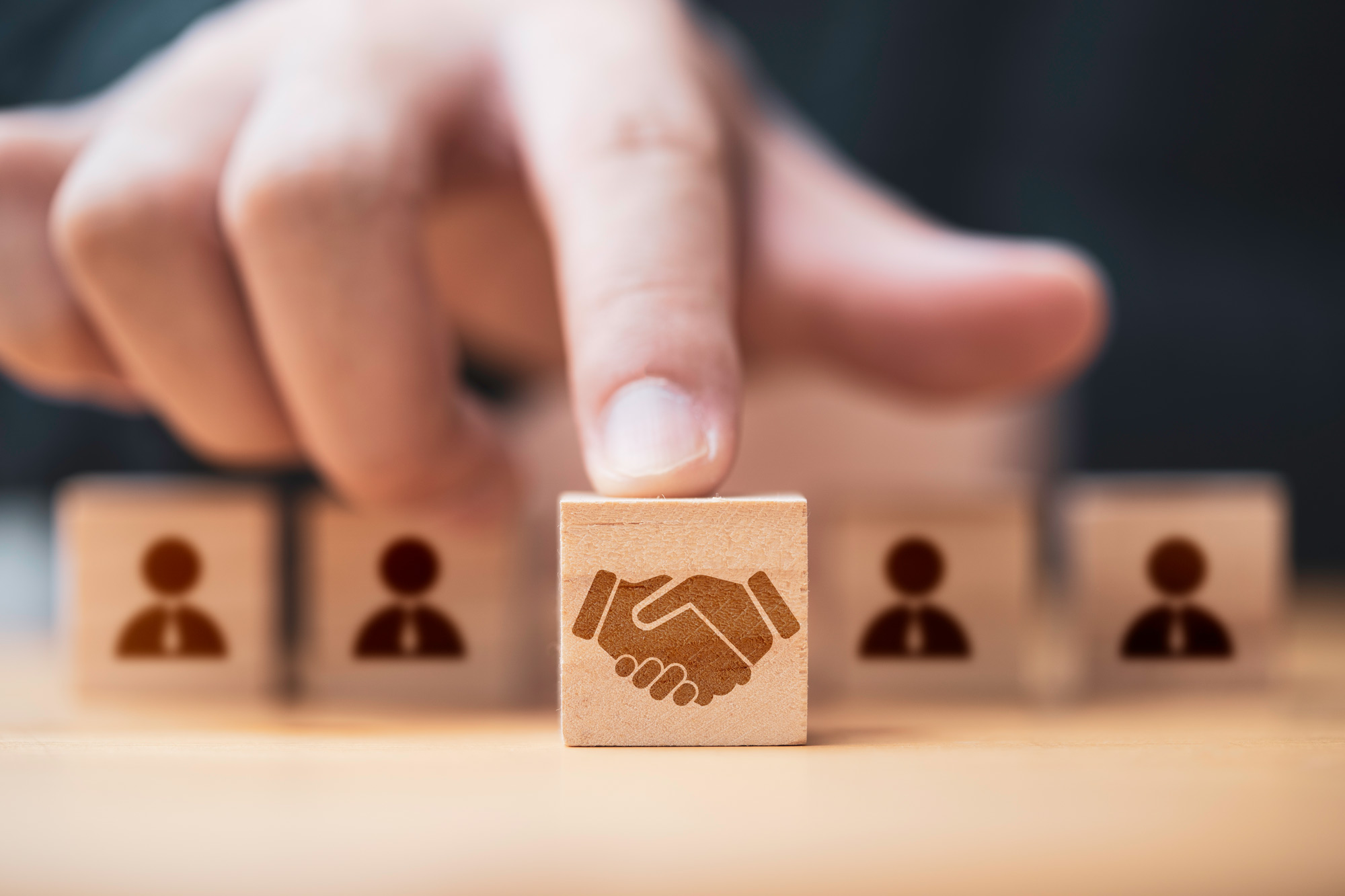 Hand setzt einen Holzwürfel mit Handschlag-Symbol zwischen Figuren – Sinnbild für Partnerschaft